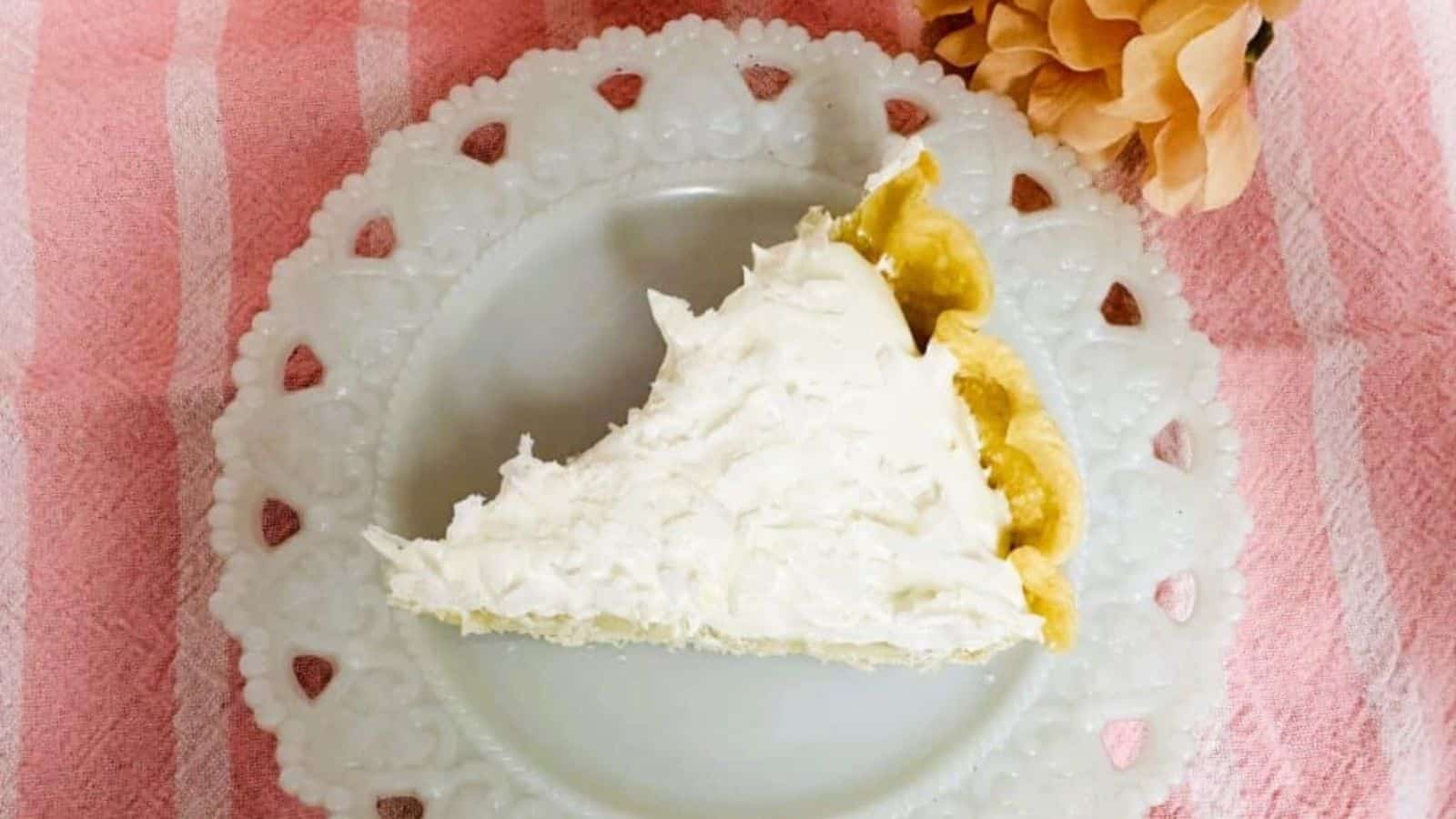 A slice of cream pie rests on a decorative white plate with a scalloped edge, set atop a pink and white striped cloth. A peach-colored flower adds charm, making this scene reminiscent of classic boomer dessert recipes that sweeten your day with nostalgia.