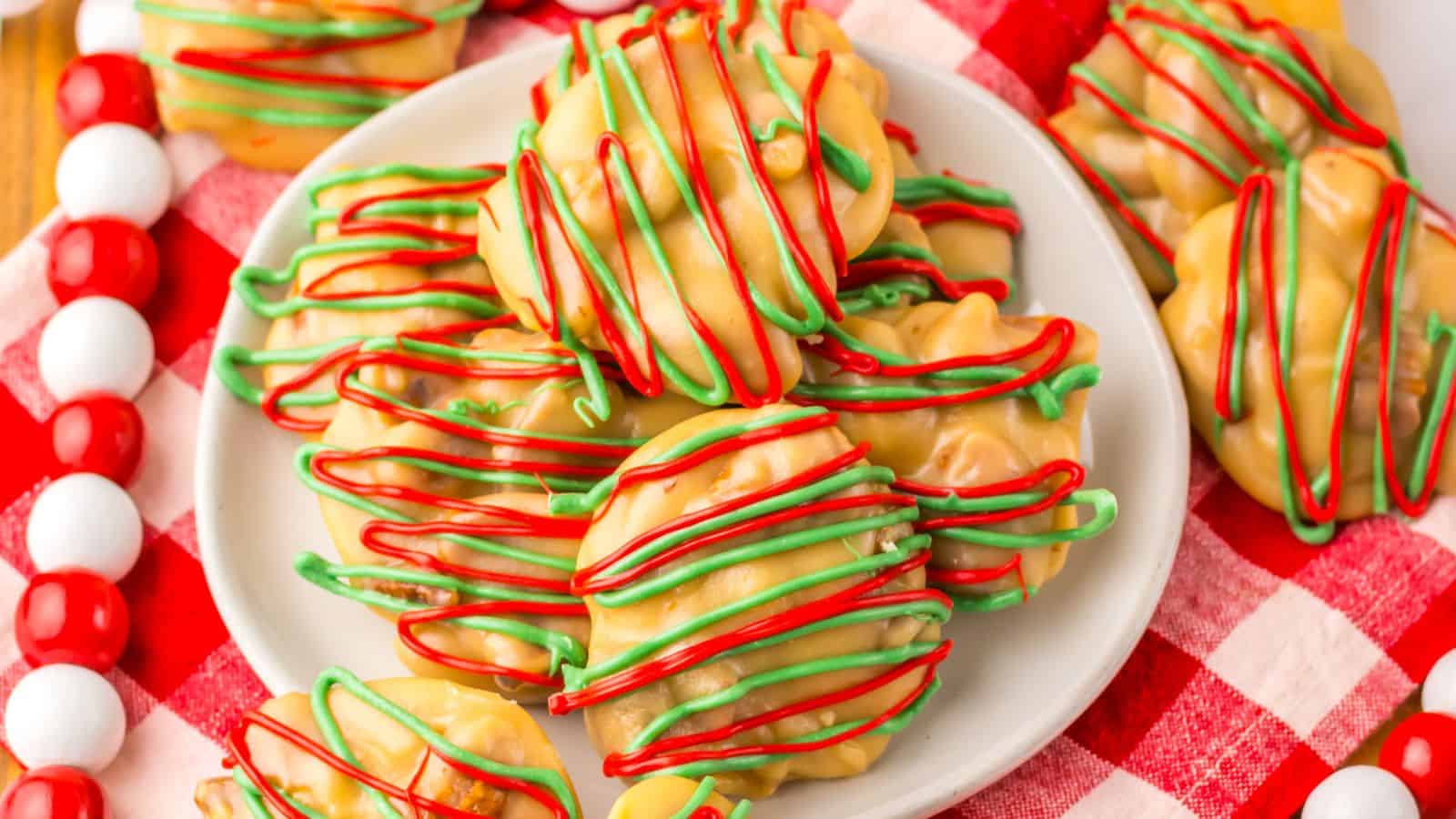 A plate of holiday-themed cookies is placed on a red and white checkered cloth. The cookies are drizzled with red and green icing over a caramel-colored base, surrounded by red and white candy beads.