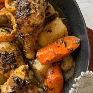 A skillet filled with flavorful roasted chicken drumsticks seasoned with herbs, surrounded by roasted potatoes, carrots, and lemon slices. This one-pan meal, set on a wooden board with a gray napkin beside it, is perfect to save time while cooking.