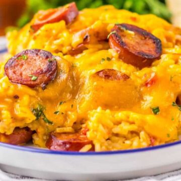 A white plate filled with a cheesy rice and sausage dish, perfect for busy weeknights. The sausage slices are mixed with the yellow cheese-covered rice, garnished with herbs. A vegetable side dish is partially visible in the background.