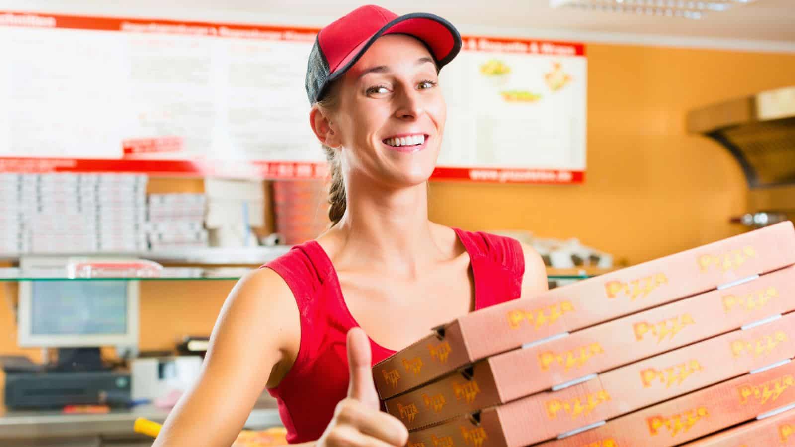 A person in a red shirt and cap holds a stack of pizza boxes while giving a thumbs-up in a restaurant setting. Menu boards and shelves are visible in the background.