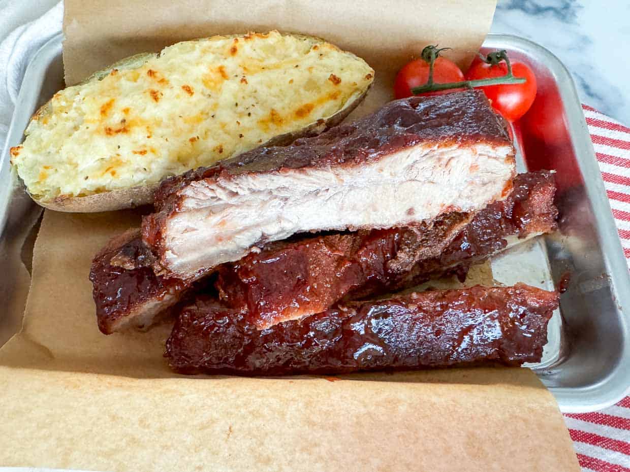 A tray with barbecue ribs, a twice-baked potato topped with cheese, and a small bunch of cherry tomatoes, all reminiscent of Grandma's recipes, placed on brown parchment paper.