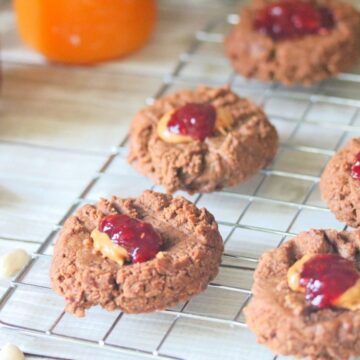 Six chocolate cookies with jelly and peanut butter centers are cooling on a wire rack. A jar of red jam, a spoon, and another jar with an orange spread are in the background on a wooden surface.