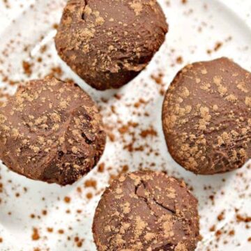 Four chocolate truffles sprinkled with cocoa powder are placed on a white, ribbed plate. Cocoa powder is scattered around the truffles on the plate.