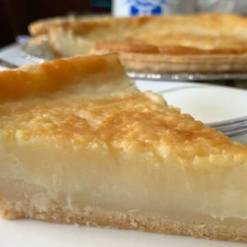 A slice of creamy pie with a golden brown crust sits on a decorative plate, a true testament to good pies. In the background, there's a larger dessert and a striped cloth. A fork is partially visible next to the slice.