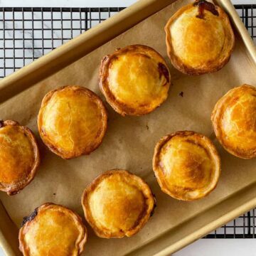A baking tray on a cooling rack holds eight round golden-brown pies, baked to perfection and evenly spaced on brown parchment paper. Their crusts contrast beautifully against the white background surface, making you wish you could enjoy one yourself.