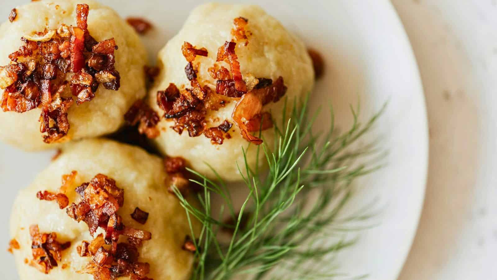 Three balls of mashed potatoes topped with crispy bacon bits are arranged on a white plate next to a sprig of fresh dill.