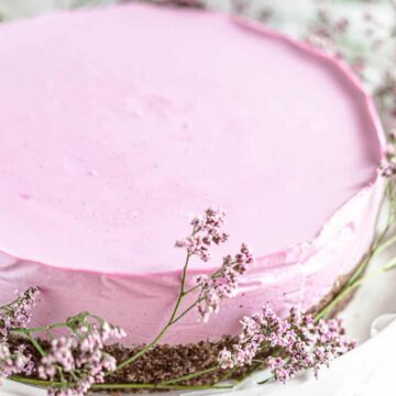 A round pink cheesecake on a white plate is garnished with small, delicate purple flowers around its base. The cheesecake has a smooth surface, with a brown crust visible at the bottom edge.