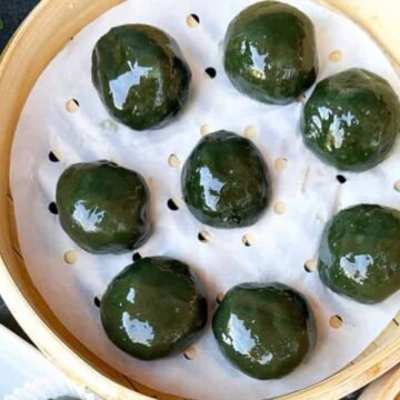 A bamboo steamer cradles eight round, glossy green dumplings on white parchment paper, inviting you to fall in love with these underrated global dishes. A small green plant peeks from the upper left corner, set against a gray surface with a wooden lid nearby.