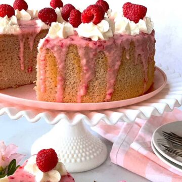 A sponge cake with pink glaze is topped with whipped cream and fresh raspberries. It is displayed on a white cake stand. There are also a few plates and forks beside the cake, and a pink napkin. A small pink flower decorates the scene.