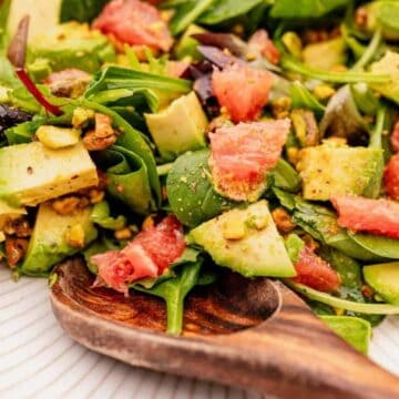 A close-up of a fresh salad featuring spinach leaves, diced avocado, grapefruit segments, and chopped pistachios. The salad is served on a white plate with a wooden serving spoon.