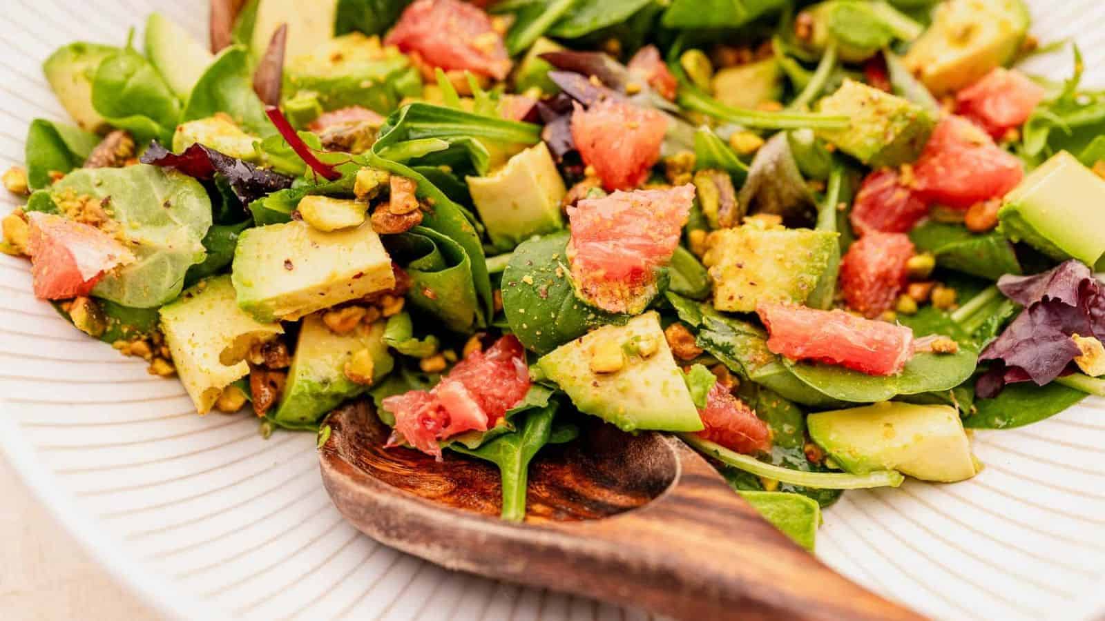 A close-up of a fresh salad featuring spinach leaves, diced avocado, grapefruit segments, and chopped pistachios. The salad is served on a white plate with a wooden serving spoon.