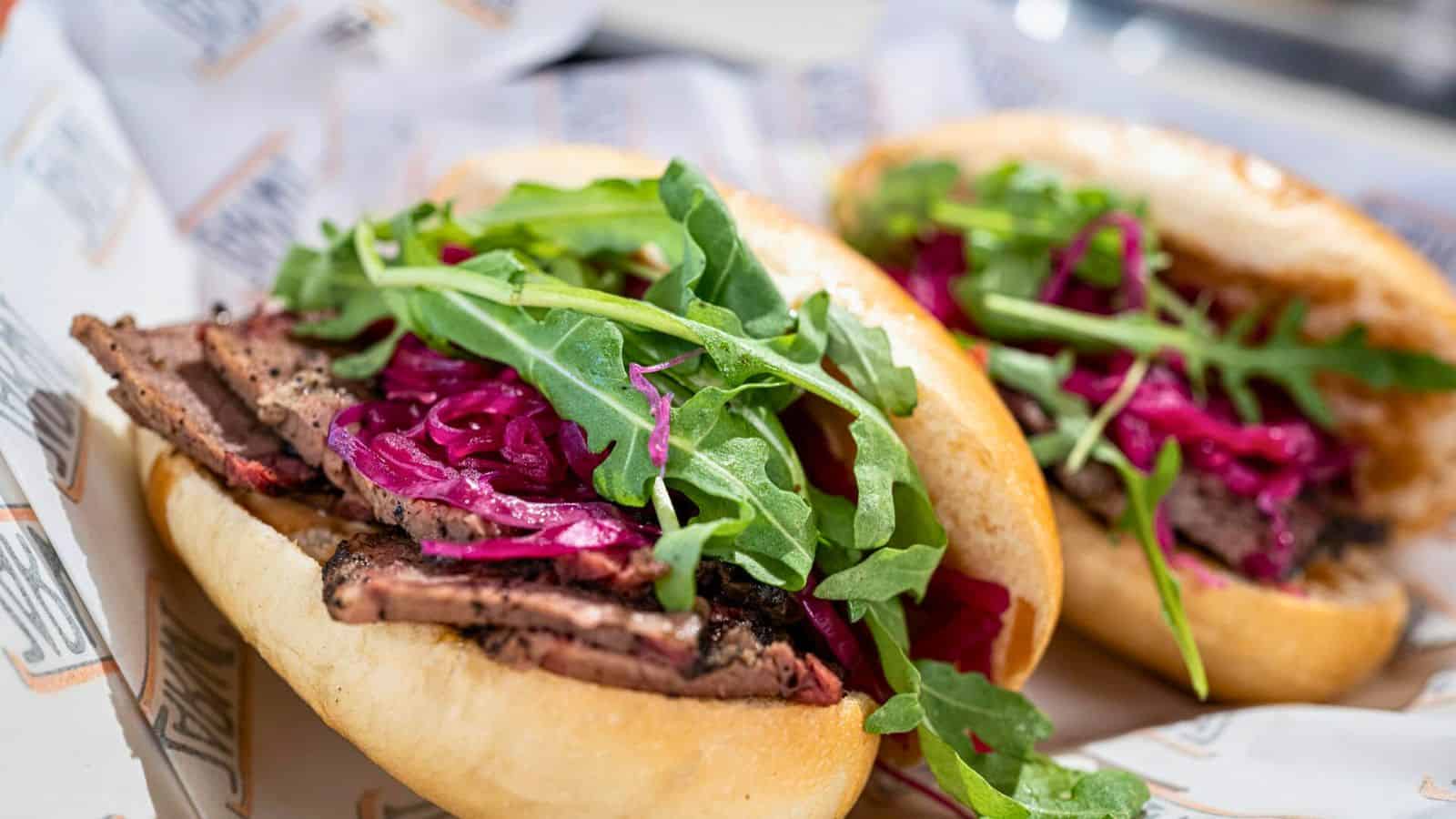 Two sandwiches filled with sliced cooked meat, purple cabbage, and fresh arugula are placed on a paper-lined surface. The bread rolls are lightly toasted.