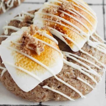 A cookie topped with two toasted marshmallows, drizzled with white icing, and sprinkled with crumbled bits. Perfect for those who love simple cookie recipes, it sits on a surface adorned with a white and black grid pattern.
