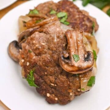 A close-up of a cooked hamburger patty topped with sliced mushrooms and chopped onions on a white plate evokes '60s recipes nostalgia. Some parsley peeks in the background on a wooden surface, ready to impress your guests with its classic charm.