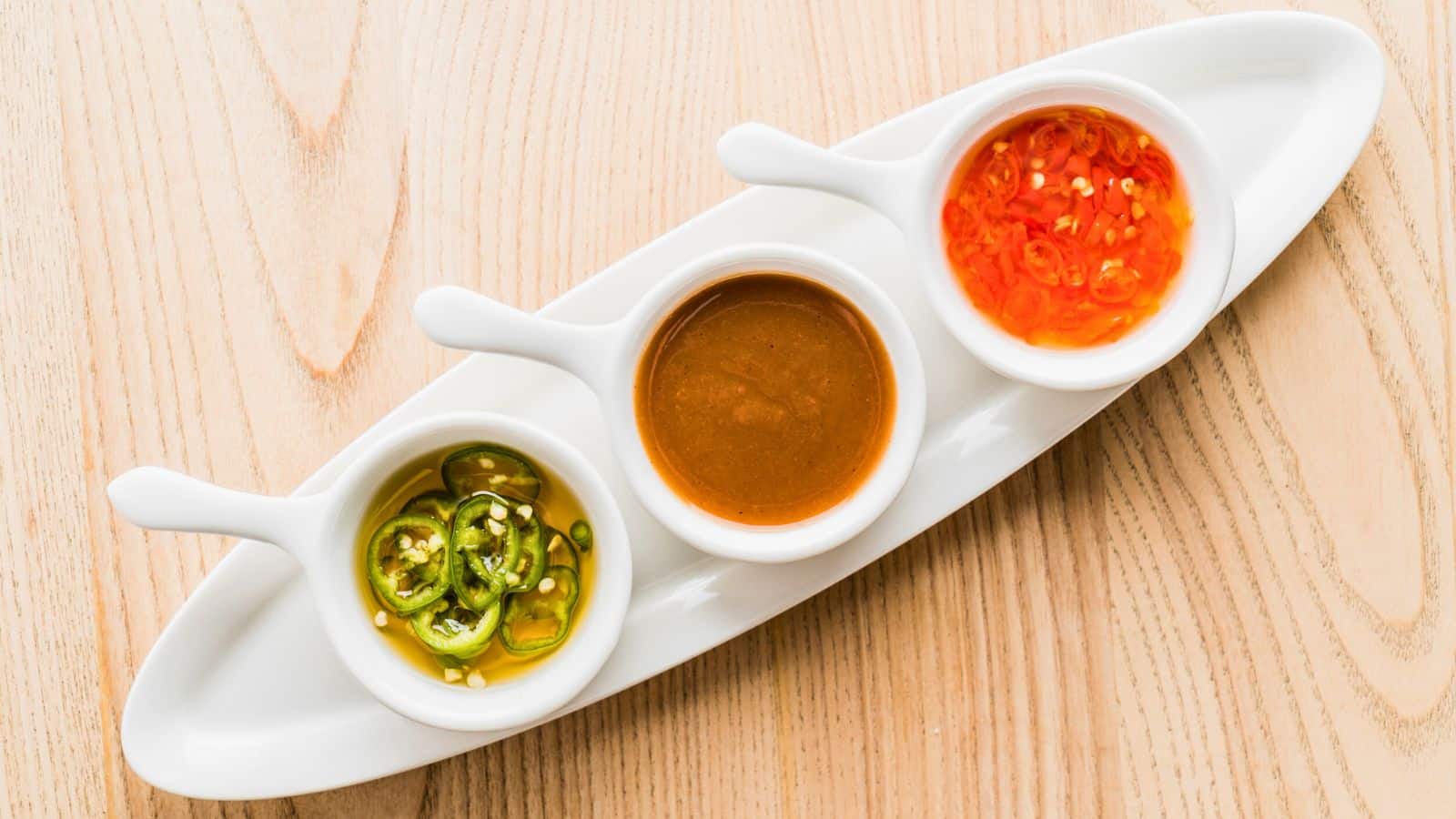 Three small white bowls sit on a long white plate, each containing a different sauce: sliced green jalapeños with seeds, a smooth brown sauce, and a red chili sauce with visible seeds. The set is placed on a light wooden surface.