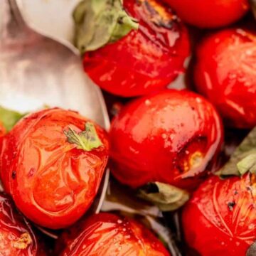 Close-up of roasted cherry tomatoes with charred spots, garnished with fresh basil leaves. A silver spoon is partially visible among the glossy tomatoes, showcasing a Mediterranean-style dish.
