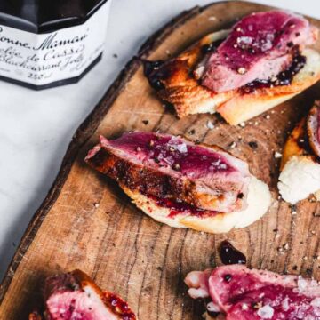 Sliced duck breast on toasted bread with berry sauce, served on a wooden board. A jar of Bonne Maman blackcurrant jelly is in the background. Salt flakes are sprinkled on top of the duck.