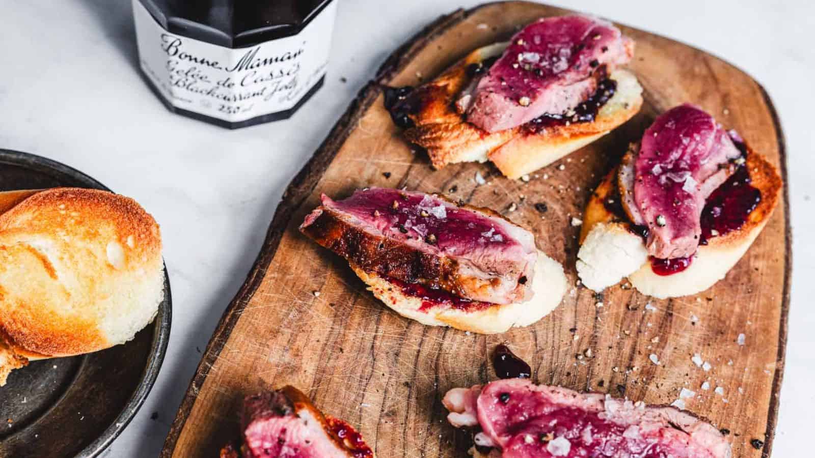 Slices of beef on toasted bread with a dark berry sauce are arranged on a wooden serving board. A jar of Bonne Maman blackcurrant jelly is nearby. Another piece of plain toasted bread is on a plate to the side.