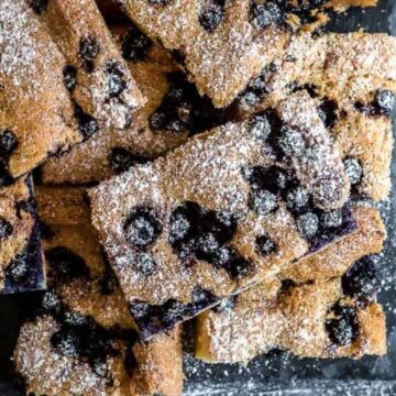 A pile of rectangular blueberry cake slices is arranged on a dark surface. These dreamy desserts are dusted with a layer of powdered sugar, revealing berries embedded in the golden-brown crumb, sure to wow any crowd.