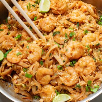 A skillet filled with shrimp pad thai, featuring noodles, shrimp, chopped peanuts, and garnished with lime wedges and herbs. Chopsticks rest on the dish.