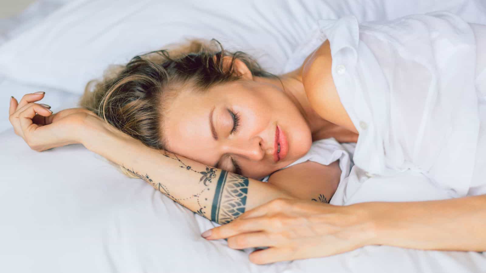 A woman with tattoos on her arm is sleeping on a bed, resting her head on a pillow. She appears relaxed, with light brown hair spread across the pillow and a white shirt partially visible. The setting is calm and peaceful.