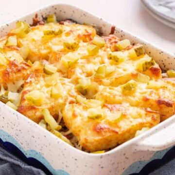 A ceramic baking dish filled with a baked casserole topped with melted cheese and diced vegetables, possibly peppers, resting on a wooden board. A grey cloth and stacked plates with a fork are in the background.