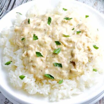 A white bowl filled with cooked white rice topped with a creamy, beige chicken and herb sauce. This kid-friendly dish is garnished with small green parsley leaves. Silver forks are visible in the background on a rustic wooden surface, tempting you to reach for seconds.