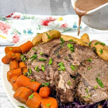 A plate of pot roast features tender beef, carrots, potatoes, and red cabbage, reminiscent of old-fashioned meals. Garnished with herbs and drizzled with sauce from a gravy boat, it sits on a floral-patterned cloth against a white tiled background.