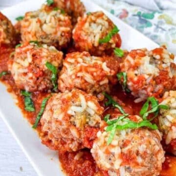 A rectangular white plate holds several meatballs covered in tomato sauce, garnished with chopped herbs. The plate rests on a light wooden table with a floral-patterned cloth partially visible, capturing the charm of Boomer Recipes—truly too good to be forgotten.