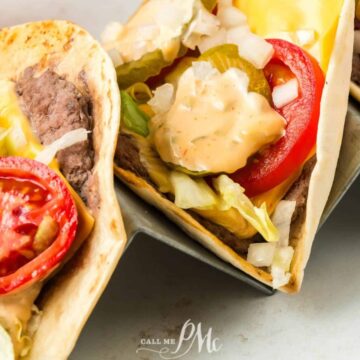 Three soft shell tacos filled with beef, sliced tomatoes, shredded lettuce, pickles, diced onions, and a creamy sauce are arranged on a metal taco holder. This next-level lunch promises to satisfy any craving. A glass with a beverage is partially visible in the background.