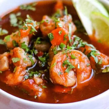 A bowl of shrimp soup garnished with cilantro. The soup has a red broth with chunks of shrimp and vegetables. A lime wedge is placed on the rim of the bowl. A spoon is partially visible beside the bowl.