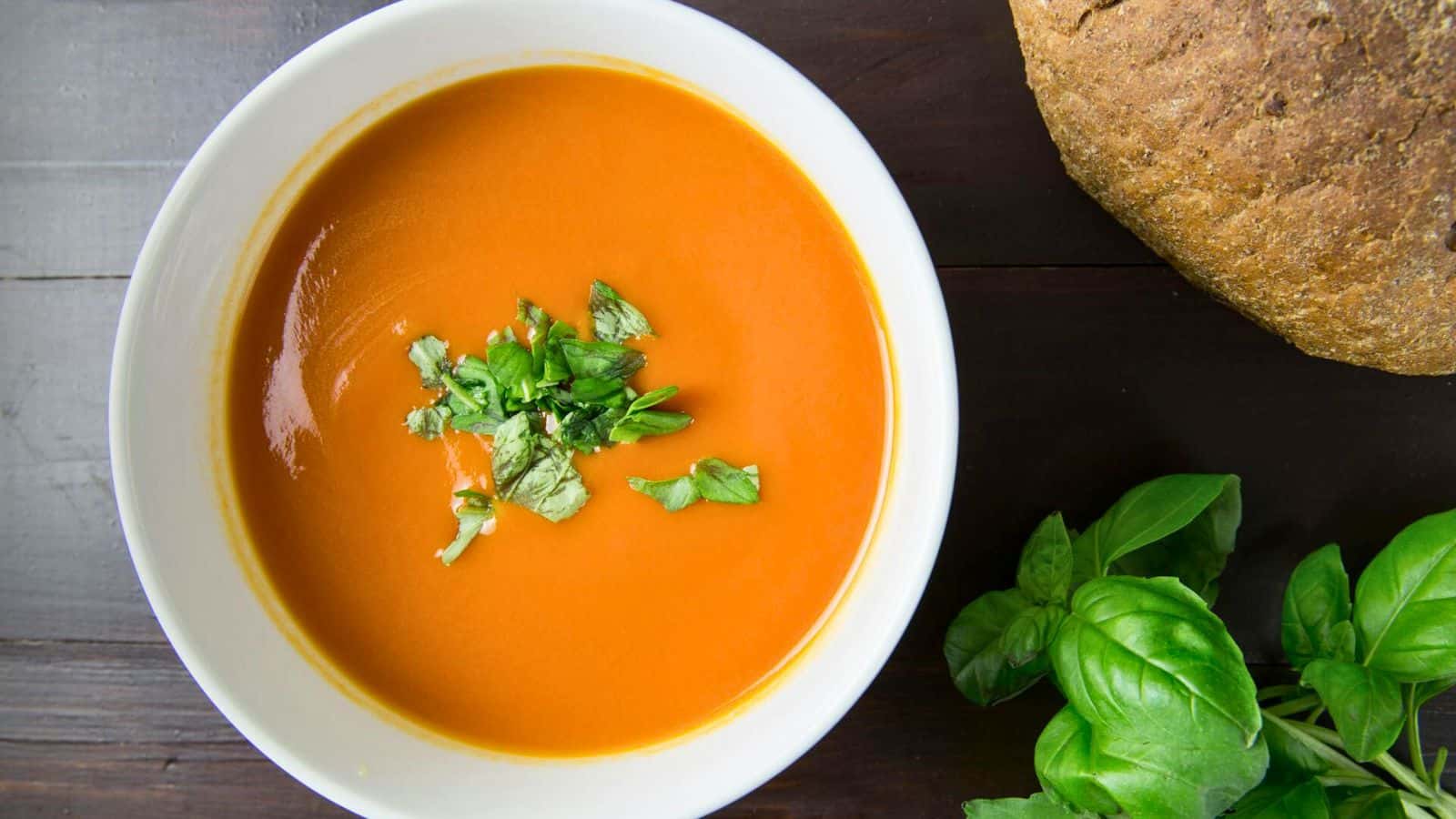 A bowl of tomato soup garnished with fresh herbs sits on a dark wooden table. A loaf of bread and fresh basil leaves are beside the bowl.