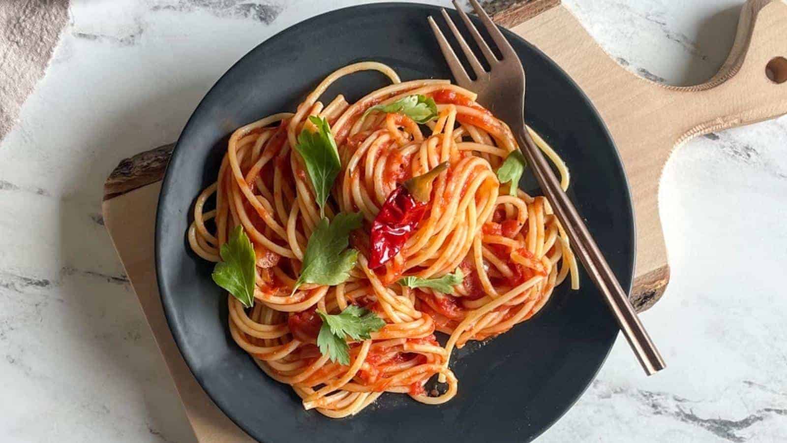 A black plate of spaghetti topped with a red tomato sauce, garnished with cilantro and red chili peppers. A metal fork rests on the wooden board beside the plate, set on a white marble surface.