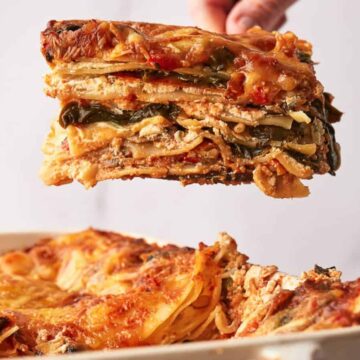 A hand holds a square piece of vegetable lasagna above a dish, showing layers of pasta, vegetables, sauce, and melted cheese. The main dish remains mostly intact in the rectangular baking pan below.