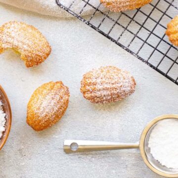 Madeleines dusted with powdered sugar are placed on a cooling rack and a light-colored surface. A wooden bowl filled with powdered sugar and a measuring cup containing more sugar are nearby. Some madeleines have bites taken out.