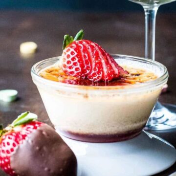 A glass dish of crème brûlée topped with sliced strawberries sits on a dark wooden table. A chocolate-covered strawberry is nearby, with scattered candy hearts. A wine glass and spoon are partially visible in the background.