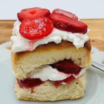 A slice of strawberry shortcake with layers of biscuit, whipped cream, and sliced strawberries on a white plate. A jar of sliced strawberries is in the background on a wooden surface.