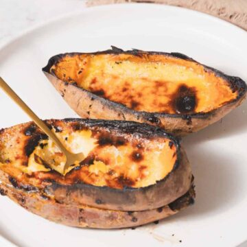 Two roasted sweet potato halves are on a white plate. Each half is charred on the edges, with a fork placed in one of them. A beige napkin is partially visible in the background.