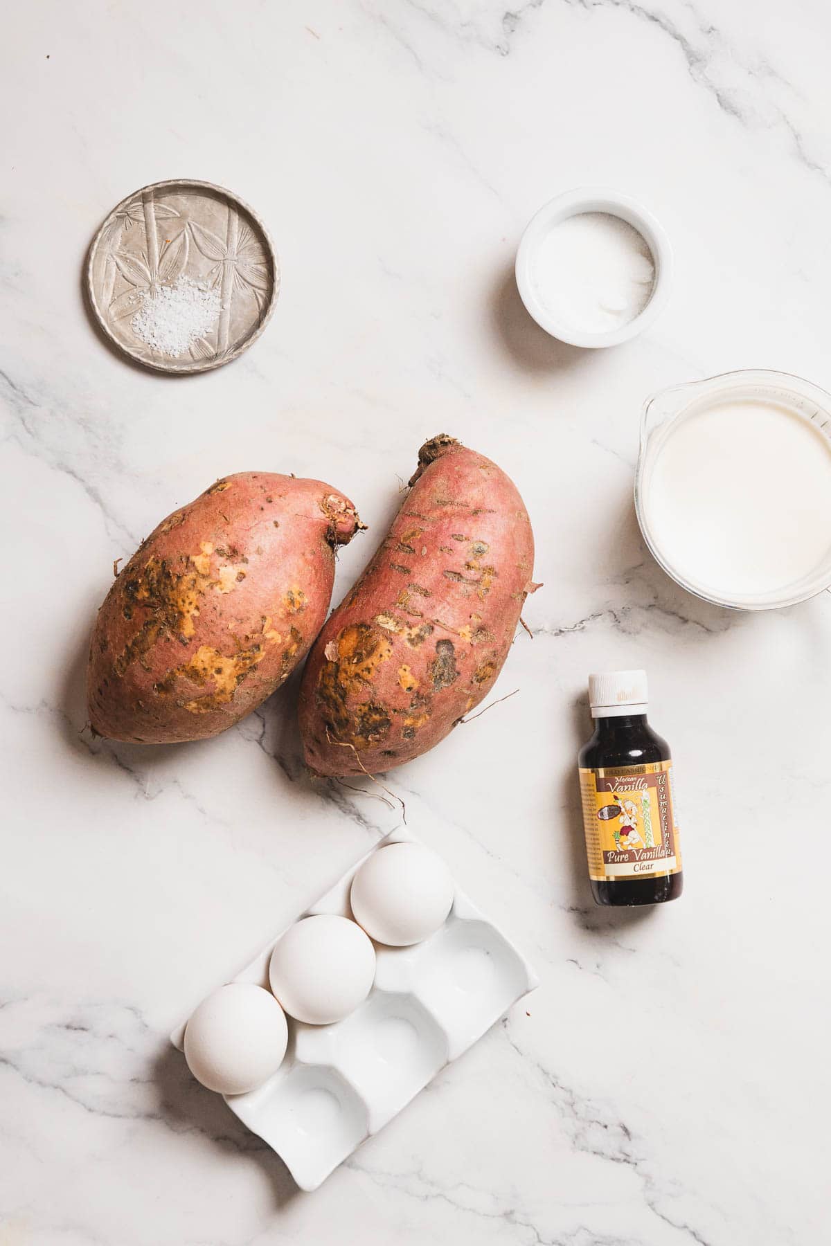 Two sweet potatoes, a small bottle of vanilla extract, three eggs in a carton, a small dish of salt, a container of sugar, and a measuring cup with milk are arranged on a marble countertop—a perfect start for a delicious sweet potato crème brûlée recipe.