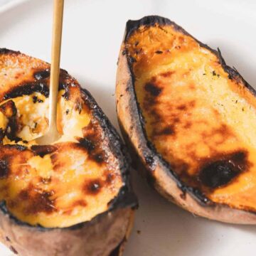 Two baked sweet potato halves on a white plate. The potatoes have a caramelized, slightly burnt surface, and one half is being scooped with a spoon.