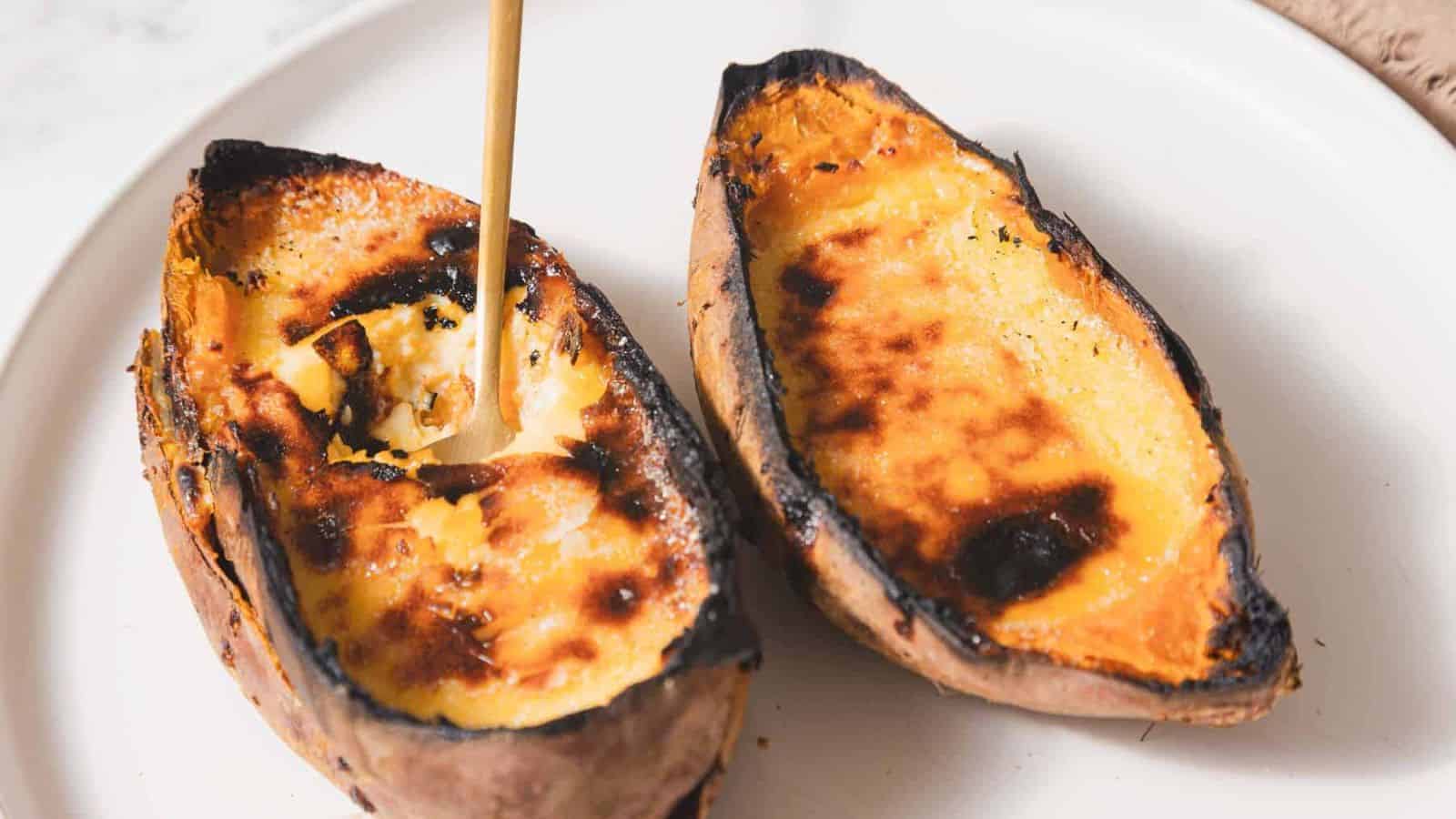 Two baked sweet potato halves on a white plate. The potatoes have a caramelized, slightly burnt surface, and one half is being scooped with a spoon.