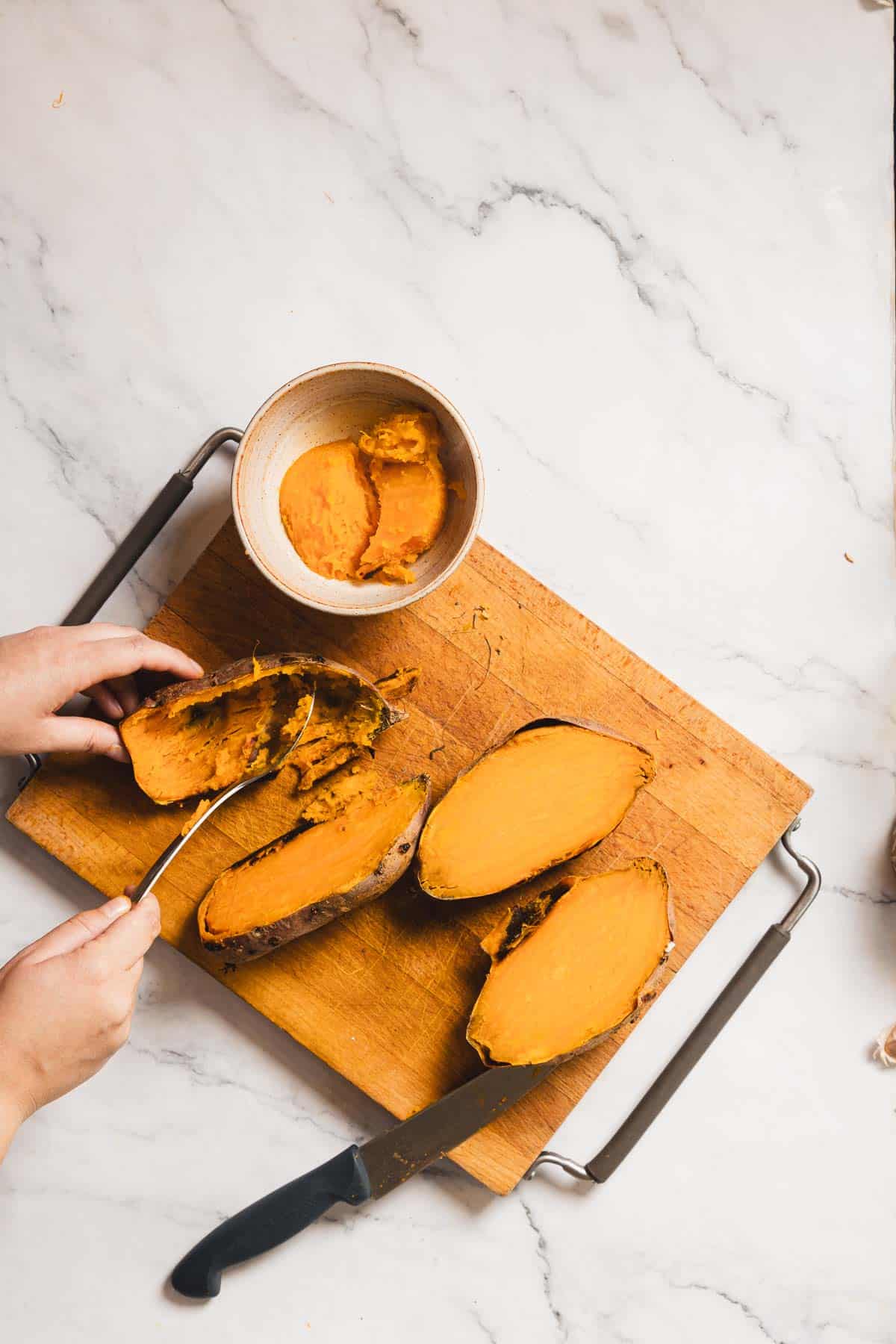 Two halves of cooked sweet potatoes sit on a wooden cutting board. A person is scooping the sweet potato flesh into a bowl with a spoon, perhaps thinking about a new best recipe. A knife rests beside them, all on a countertop with a marble-like surface.