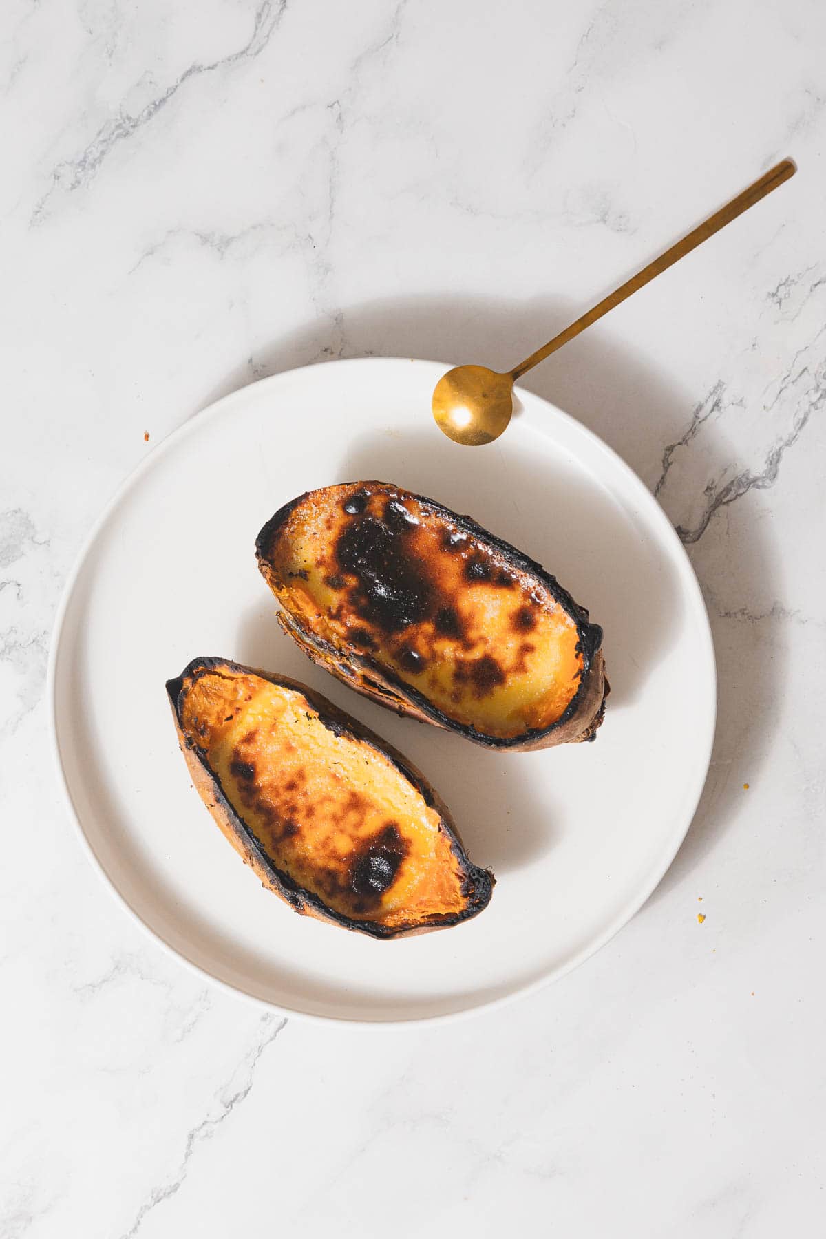 Two pieces of charred toast sit on a white plate with a golden spoon beside them, as if waiting for a spread of Sweet Potato Creme Brûlée. The plate rests on a marble surface. The toast appears crisp and browned, suggesting it may have been overcooked.