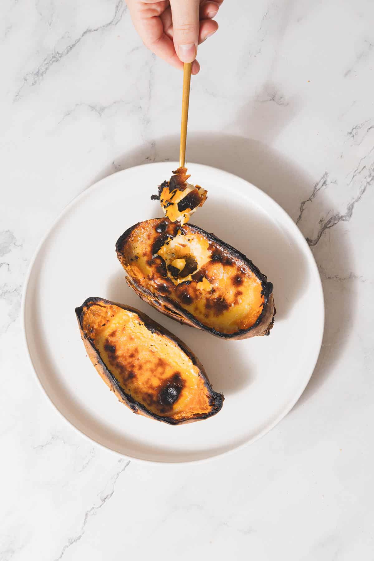 A hand holds a spoon above a white plate with two charred Sweet Potato Creme Brûlée desserts, elegantly served in caramelized squash halves. The background showcases a sleek marble surface.