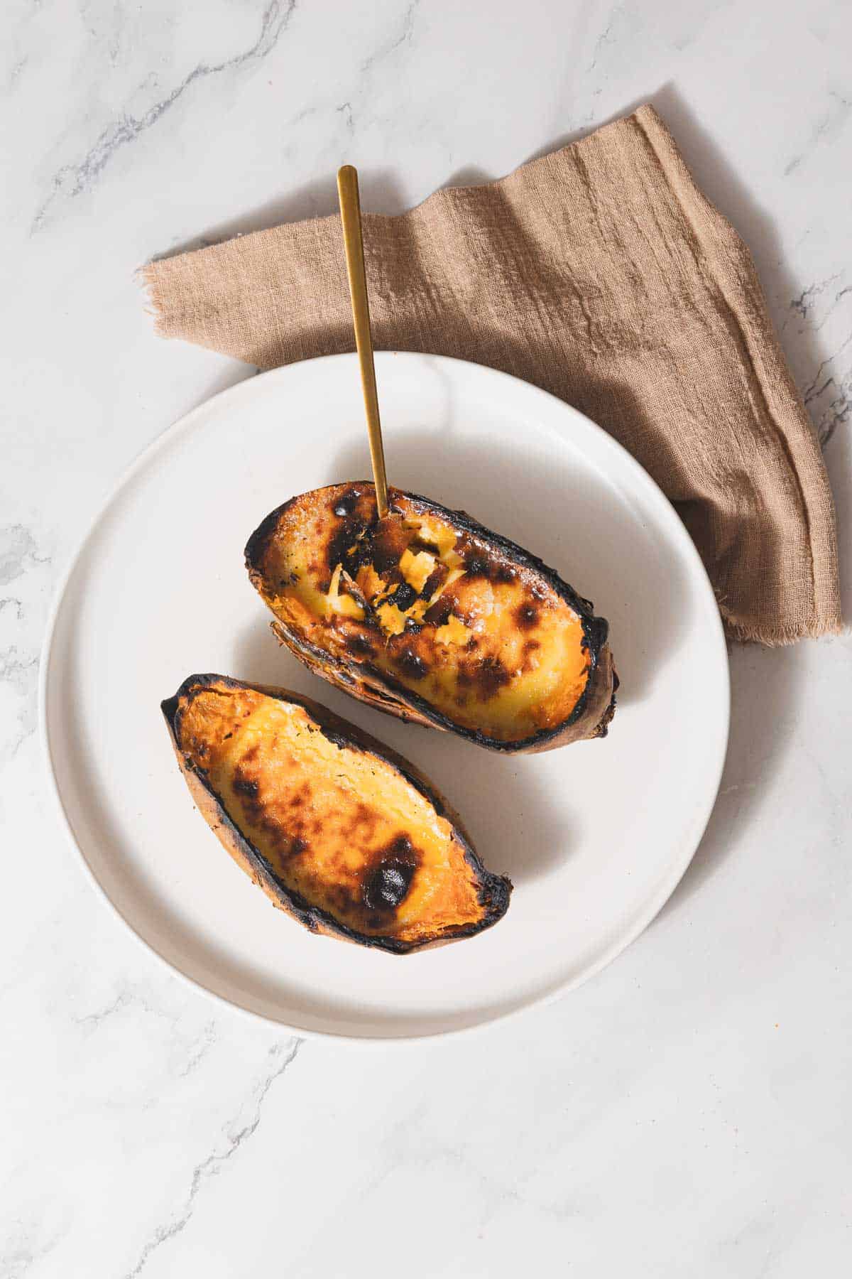 A white plate holds two halves of a roasted acorn squash with charred edges, reminiscent of a sweet potato dish, filled with cubes and a glossy brown glaze. A gold spoon rests elegantly on the plate, while a brown napkin lies nearby on the cool marble surface.