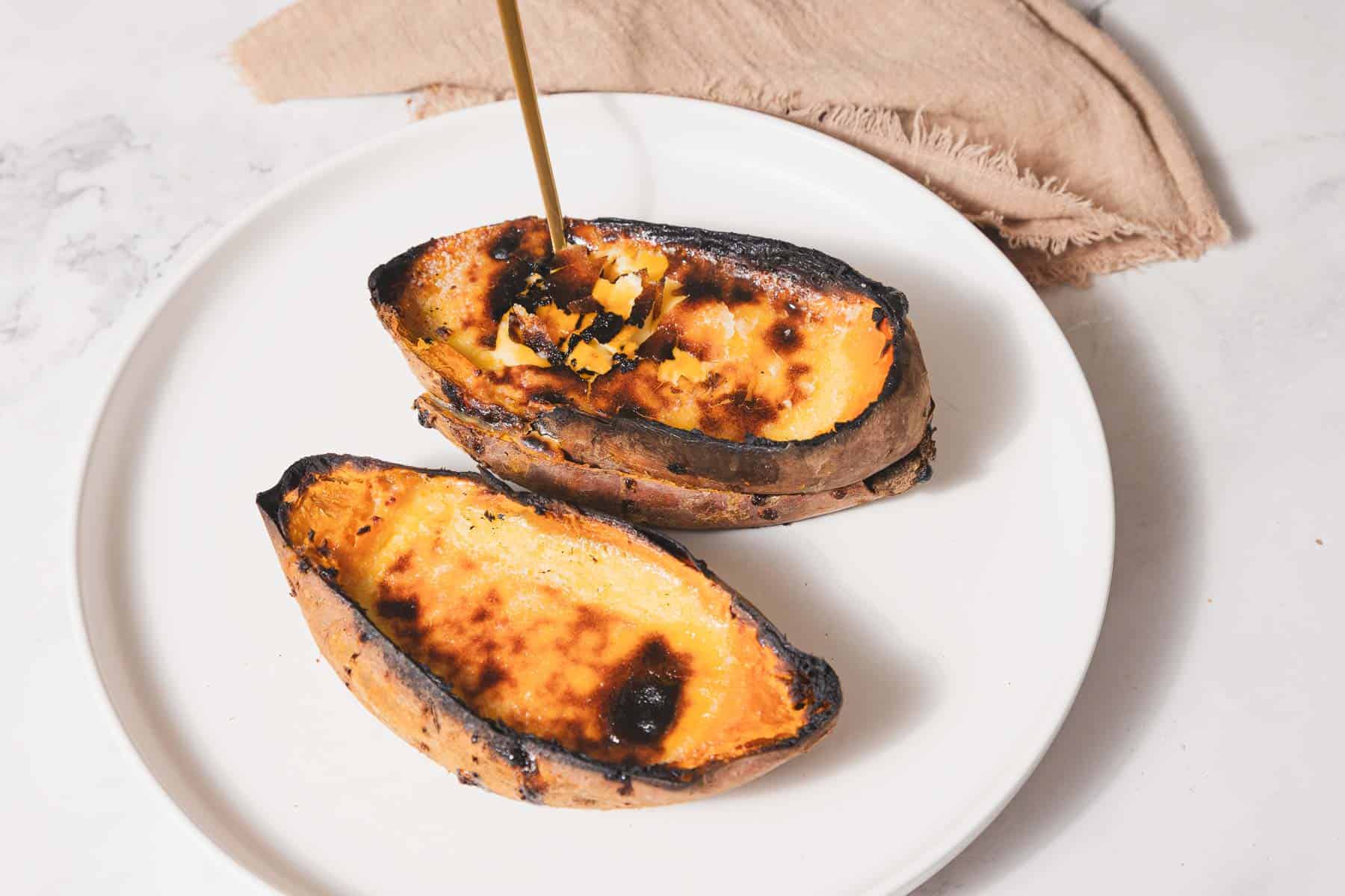 A white plate holds two halved, roasted sweet potatoes with charred edges, reminiscent of creme brûlée. A drizzle of syrup is being poured onto them, adding a final touch to this delightful recipe. A beige napkin is visible in the background on the marble surface.