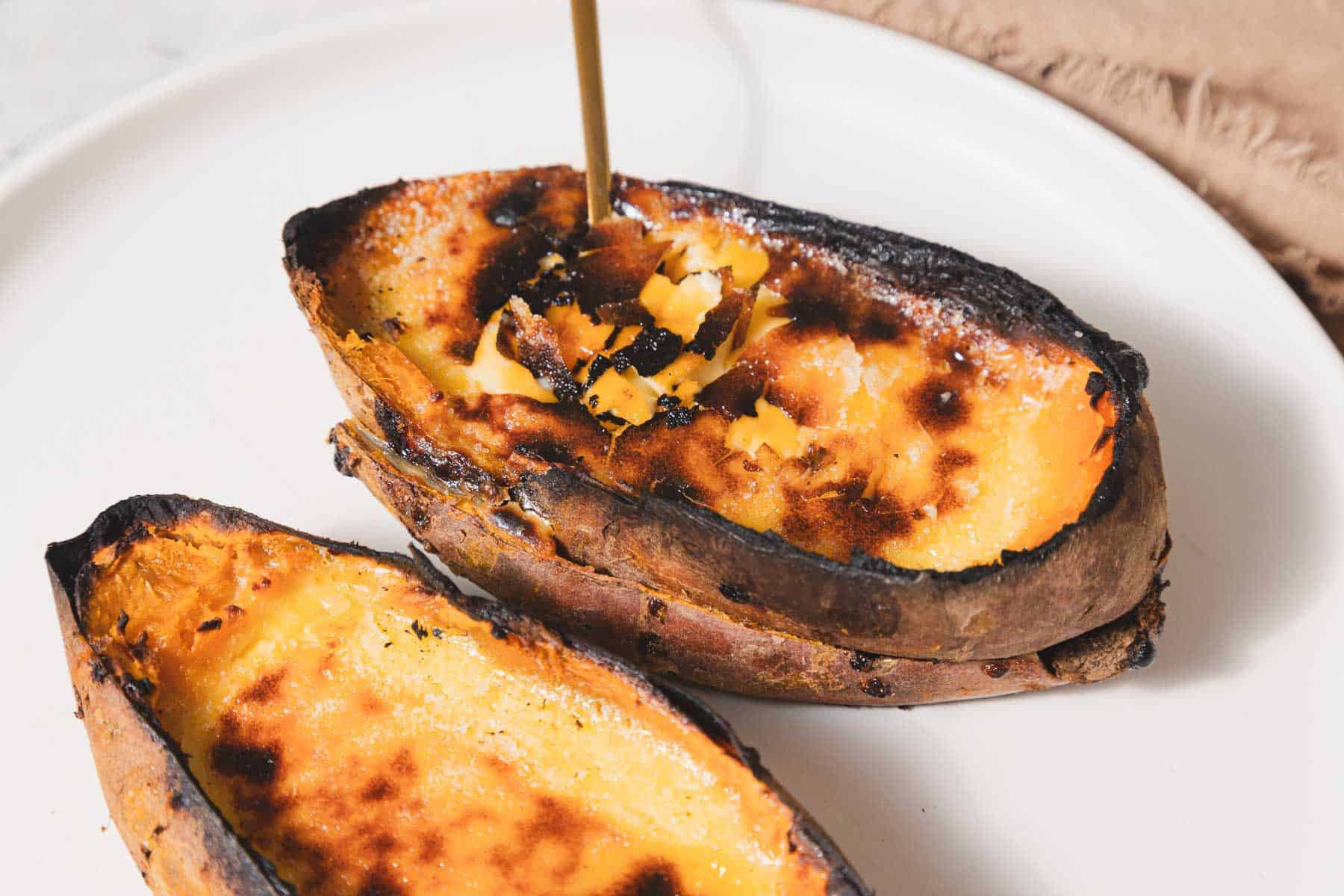 Two halves of roasted sweet potatoes with caramelized tops sit on a white plate, resembling the delicious crust of a creme brûlée. A drizzle of honey is being poured onto one half, adding to its sweet allure. A brown napkin is partially visible in the background.