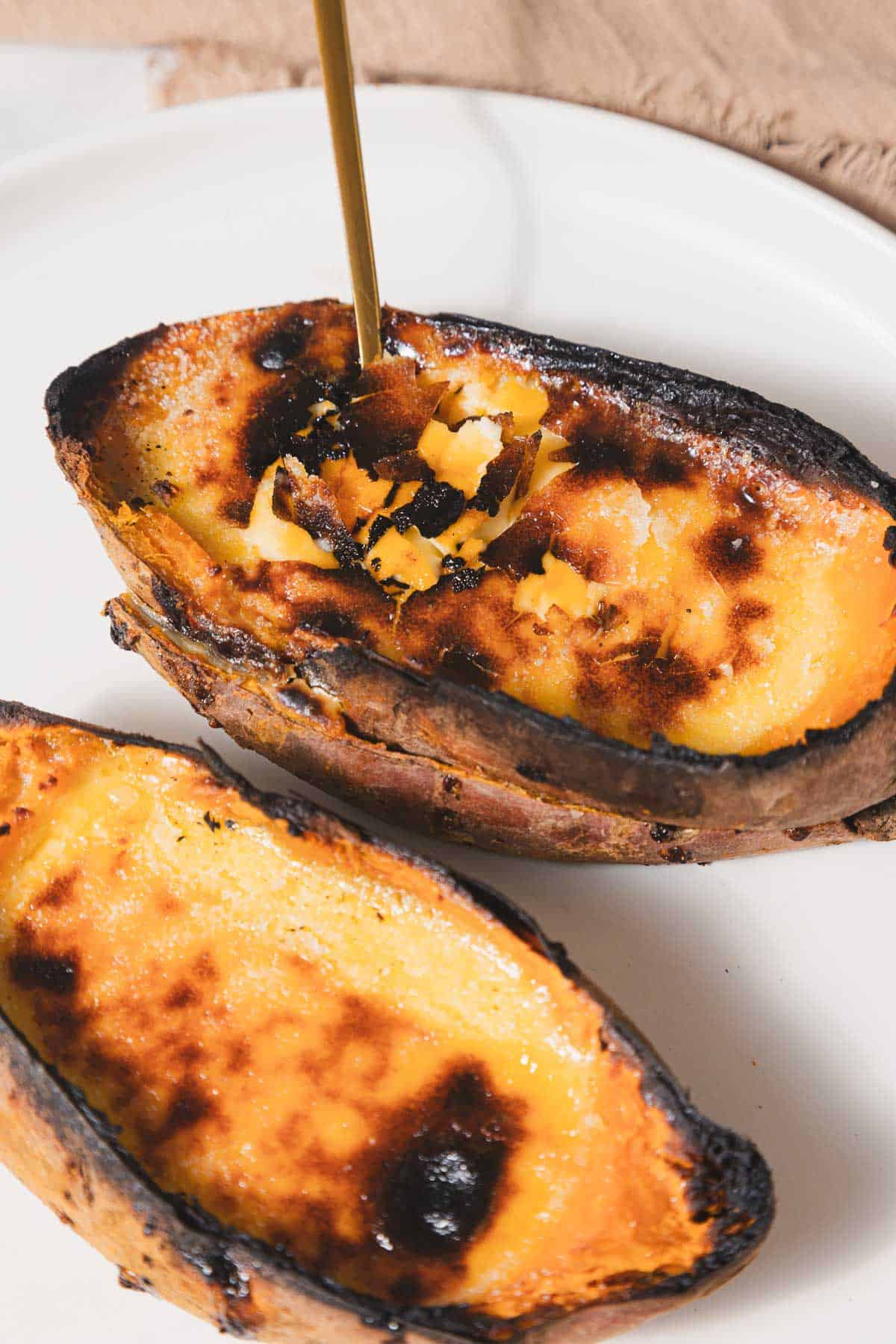 Two halved sweet potatoes, reminiscent of a sweet potato crème brûlée, with caramelized tops rest on a white plate. A spoon pierces one, revealing its smooth, creamy texture. The slightly charred skin contrasts beautifully with the rich orange interior—truly the best recipe for indulgence.