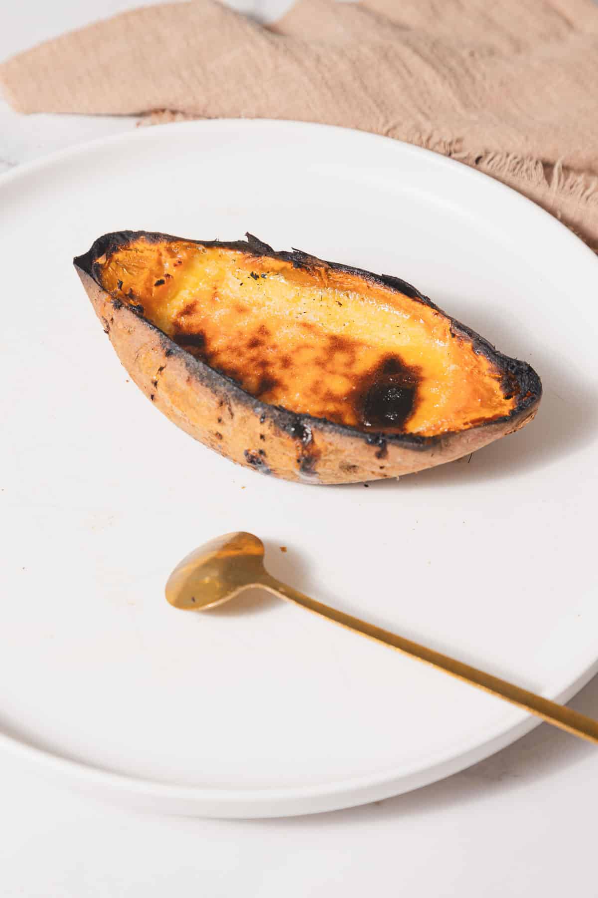 A half of a baked sweet potato with a browned top is placed on a white plate, reminiscent of a Creme Brûlée recipe. A gold spoon rests nearby, ready to dive in. A beige cloth is in the background.